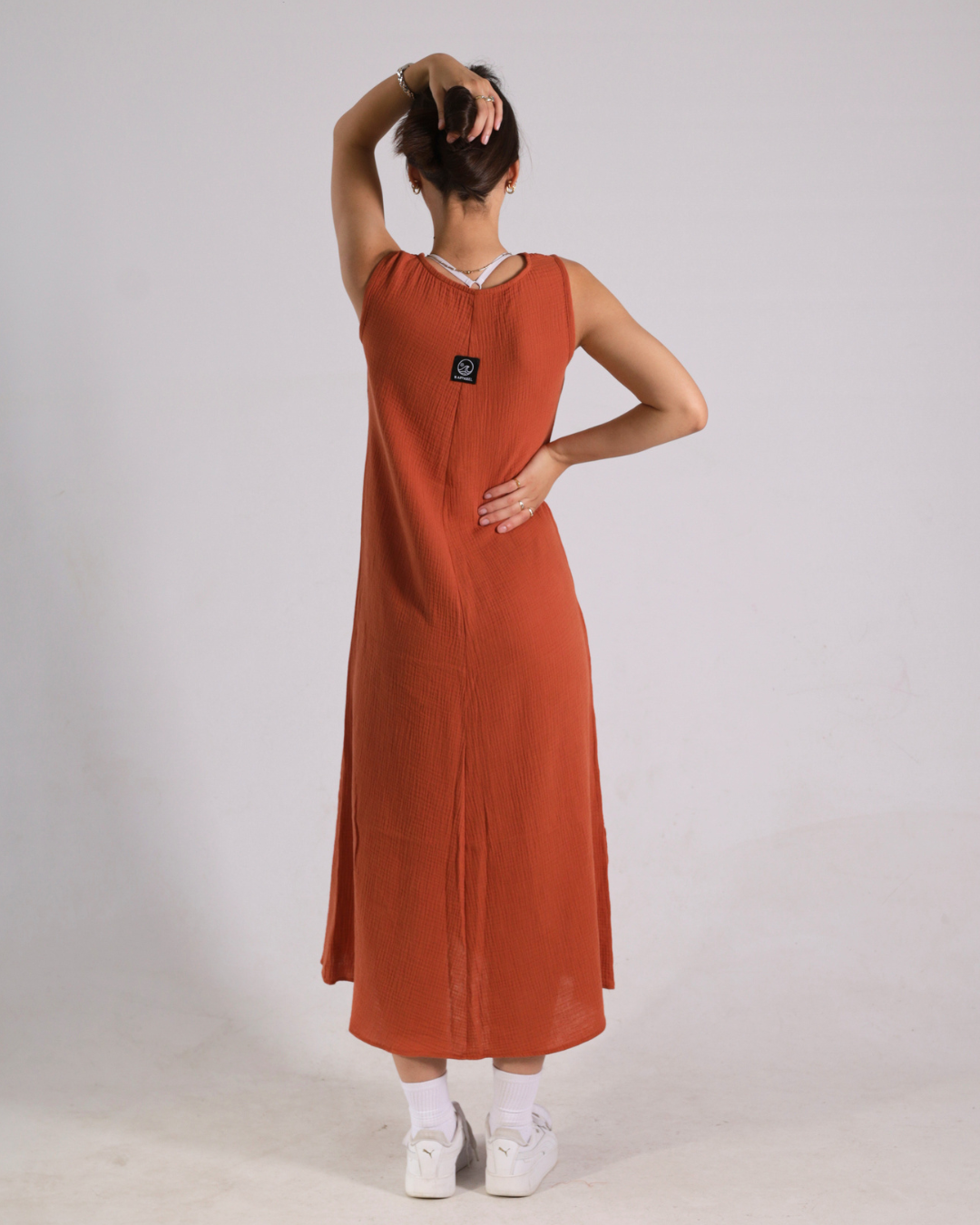 A woman shown from the back wearing a sleeveless rust-orange maxi dress with a small black square patch on the upper back, paired with white socks and white sneakers, against a light gray studio background.