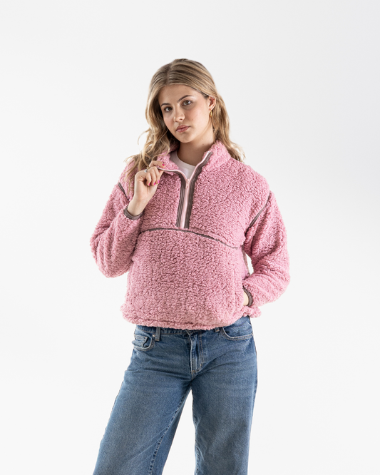 A woman modeling a pink sherpa fleece quarter-zip pullover jacket with a brown zipper trim, paired with blue jeans, against a plain white studio background.