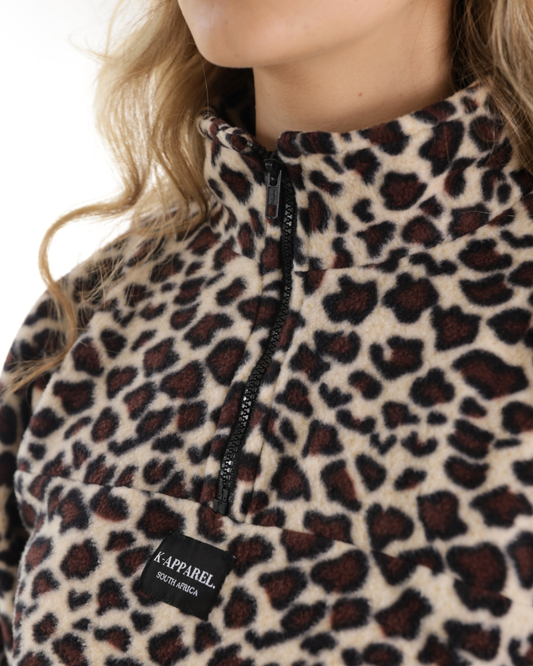 Close-up of a person wearing a leopard-print fleece half-zip pullover. The garment has a beige base with dark brown and black leopard spots, a black zipper, and a black brand patch reading K-APPAREL SCROUDDERS.