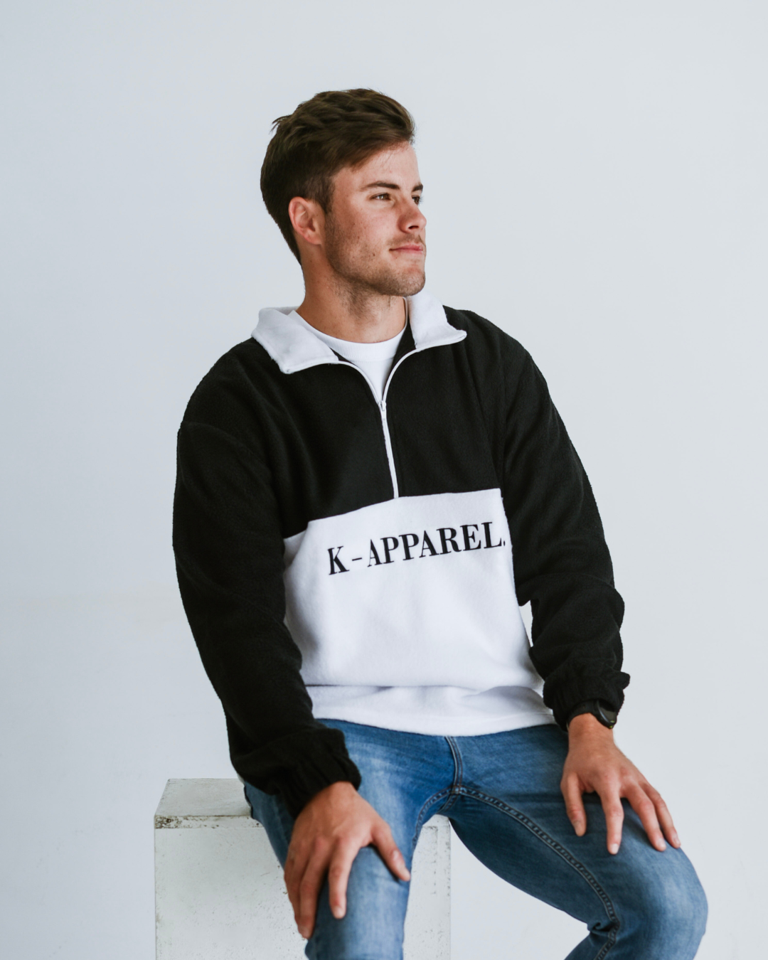 Man wearing a black and white retro fleece quarter zip K-Apparel pullover , sitting on a white stool against a light gray background