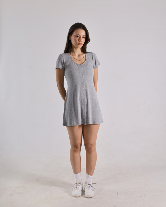 A woman modeling a light gray short-sleeve button-front mini dress with white socks and white sneakers, standing against a plain light gray studio background.