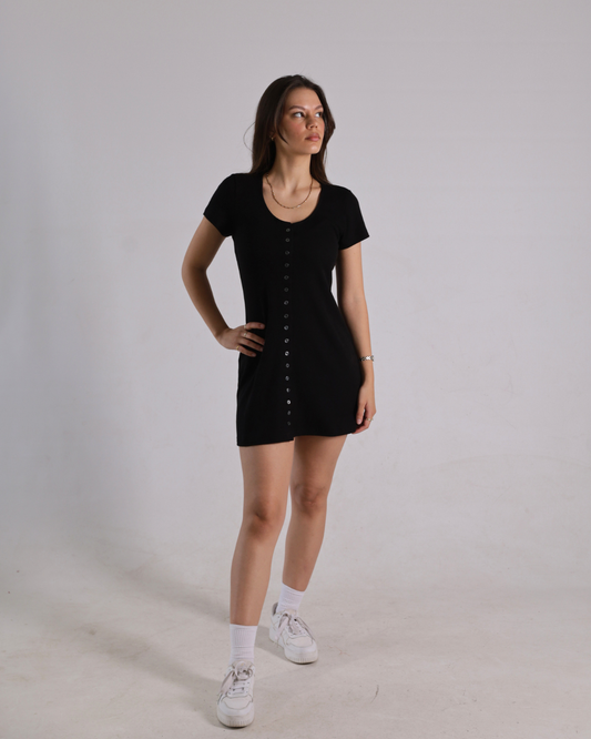 A woman modeling a short-sleeve black mini dress with a button-down front, paired with white socks and white sneakers, standing against a plain light gray studio background.