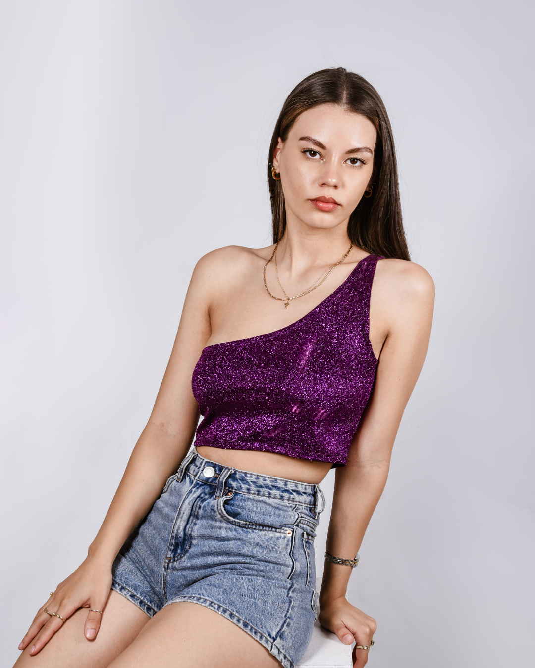 Woman wearing a purple glittery top and denim shorts against a light gray background