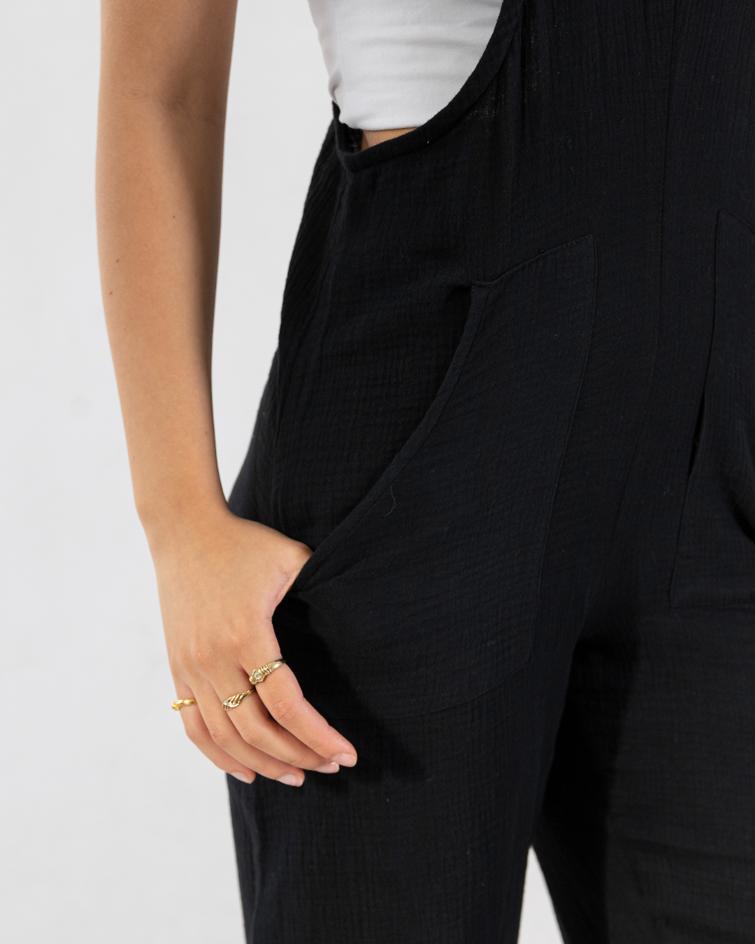 Close-up of a person wearing black textured overall-style pants with a white top, showing the side pocket and hand resting in the pocket against a light gray background.