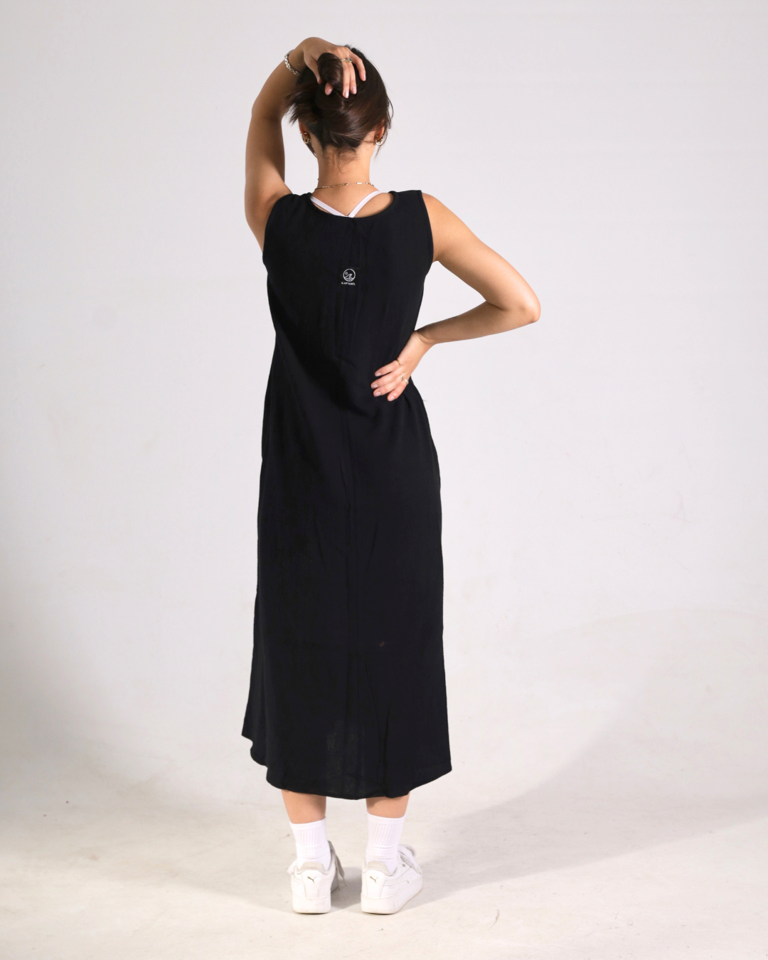 A woman wearing a sleeveless black midi dress, viewed from the back. She has her hair in a bun, wearing white socks and white platform sneakers, against a plain light gray studio background.
