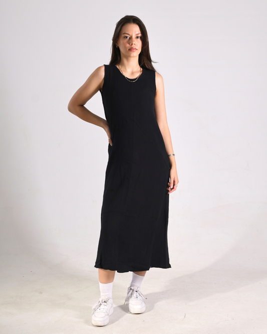 A woman modeling a sleeveless black midi dress with side pockets, paired with white socks and white sneakers, standing against a plain light gray studio background.