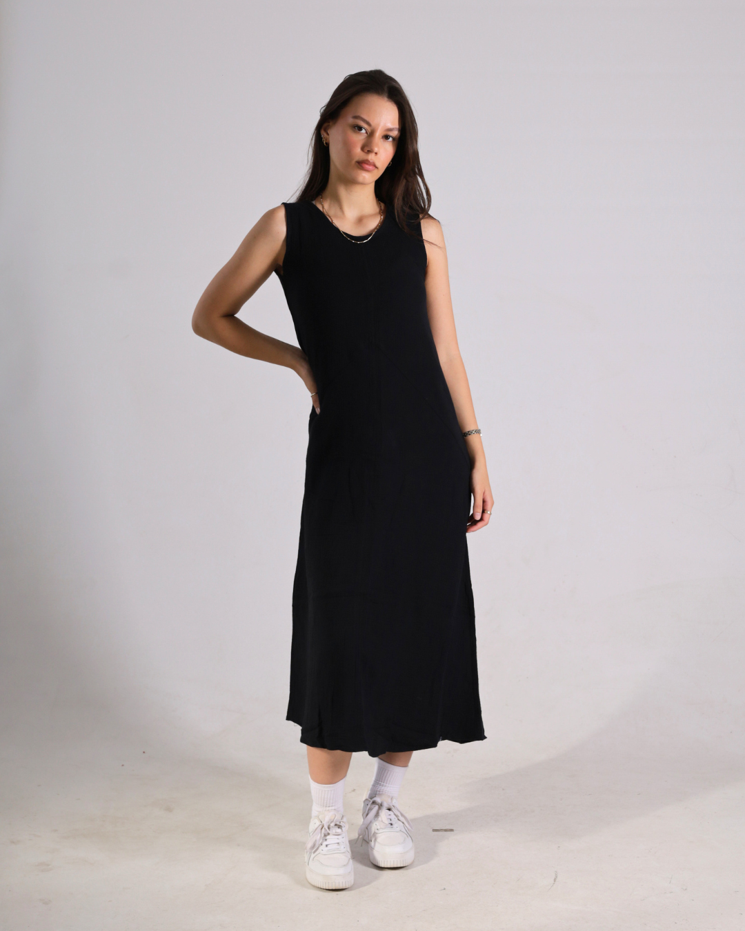 A woman modeling a sleeveless black midi dress with a scoop neckline, paired with white socks and white sneakers, standing against a plain light gray studio background.