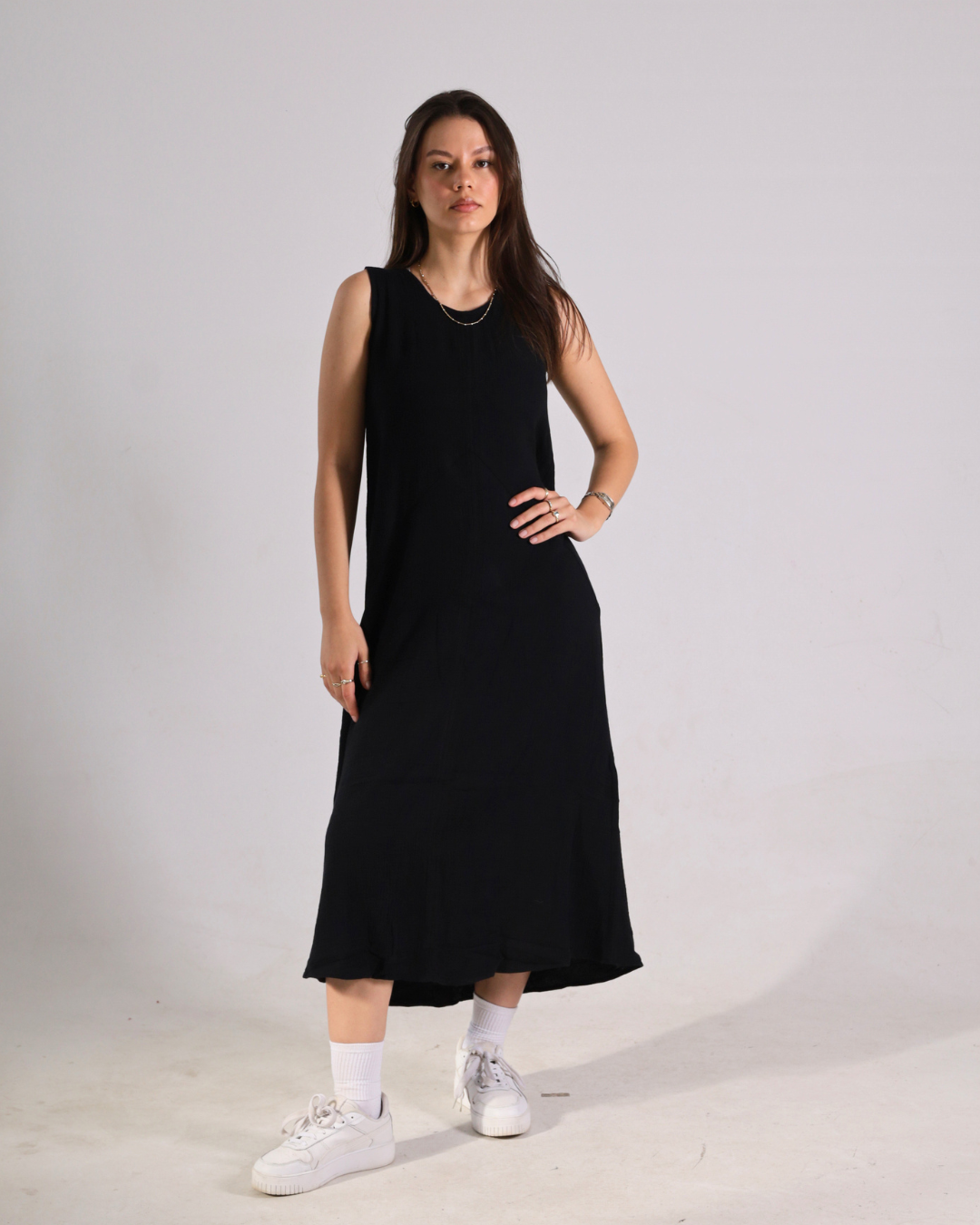 A woman modeling a sleeveless black midi dress with white crew socks and white sneakers, standing against a plain light gray studio background.