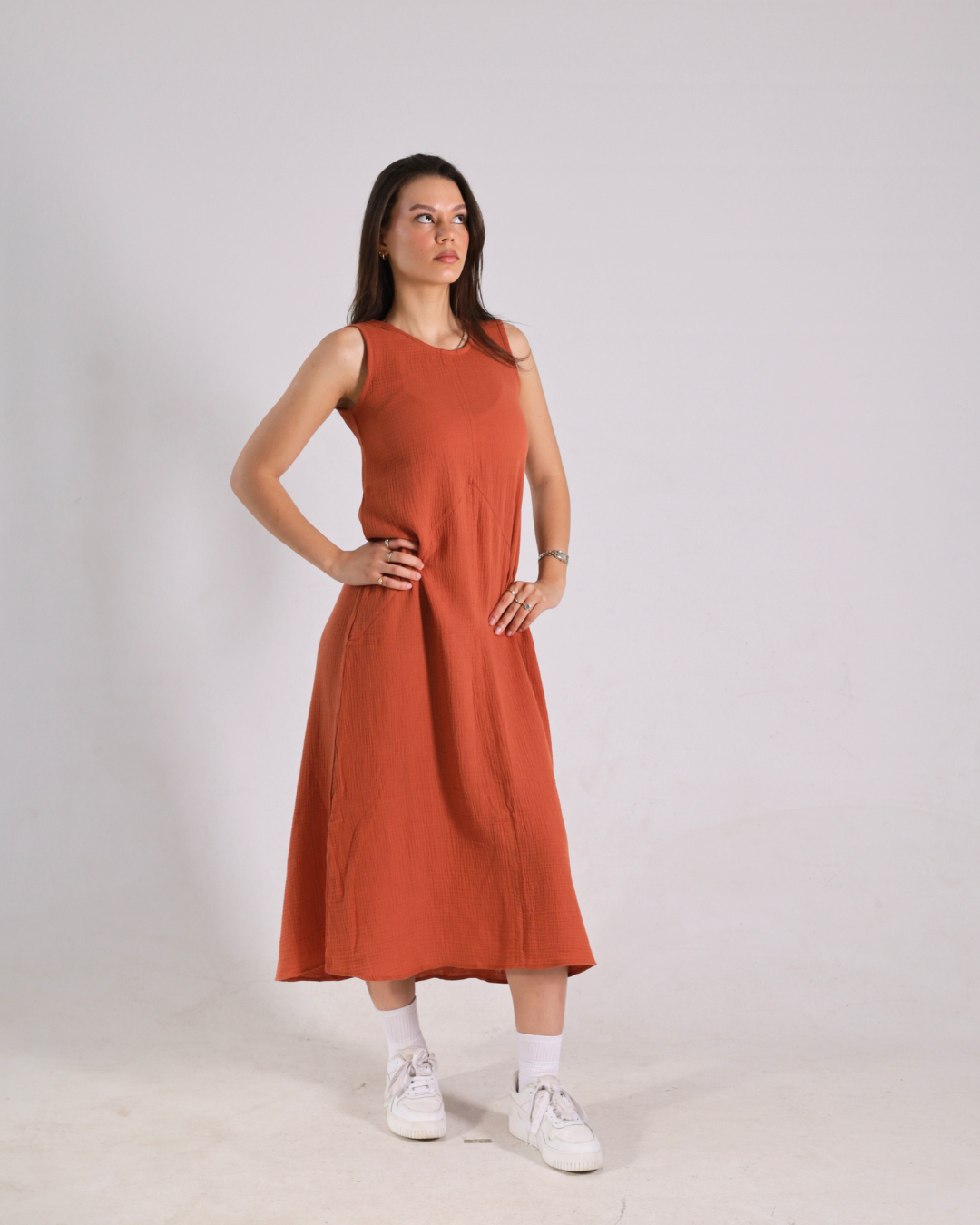 A woman modeling a sleeveless rust-orange midi dress with side pockets, paired with white socks and sneakers, standing against a light gray studio background.