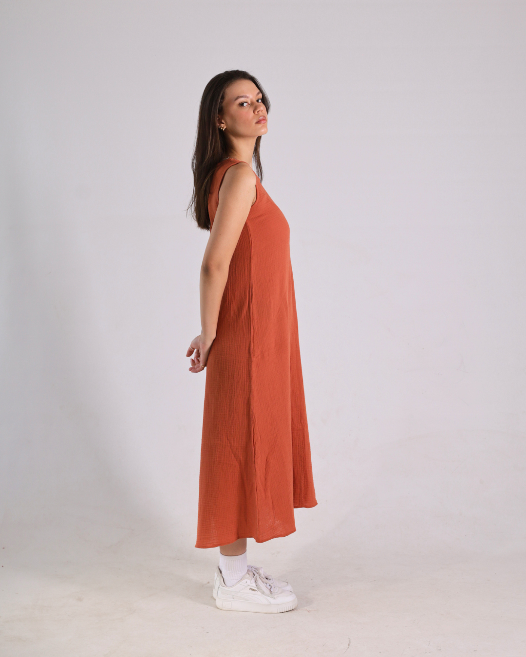 A woman modeling a sleeveless, rust-orange midi dress in a side-profile view against a plain light gray studio background.