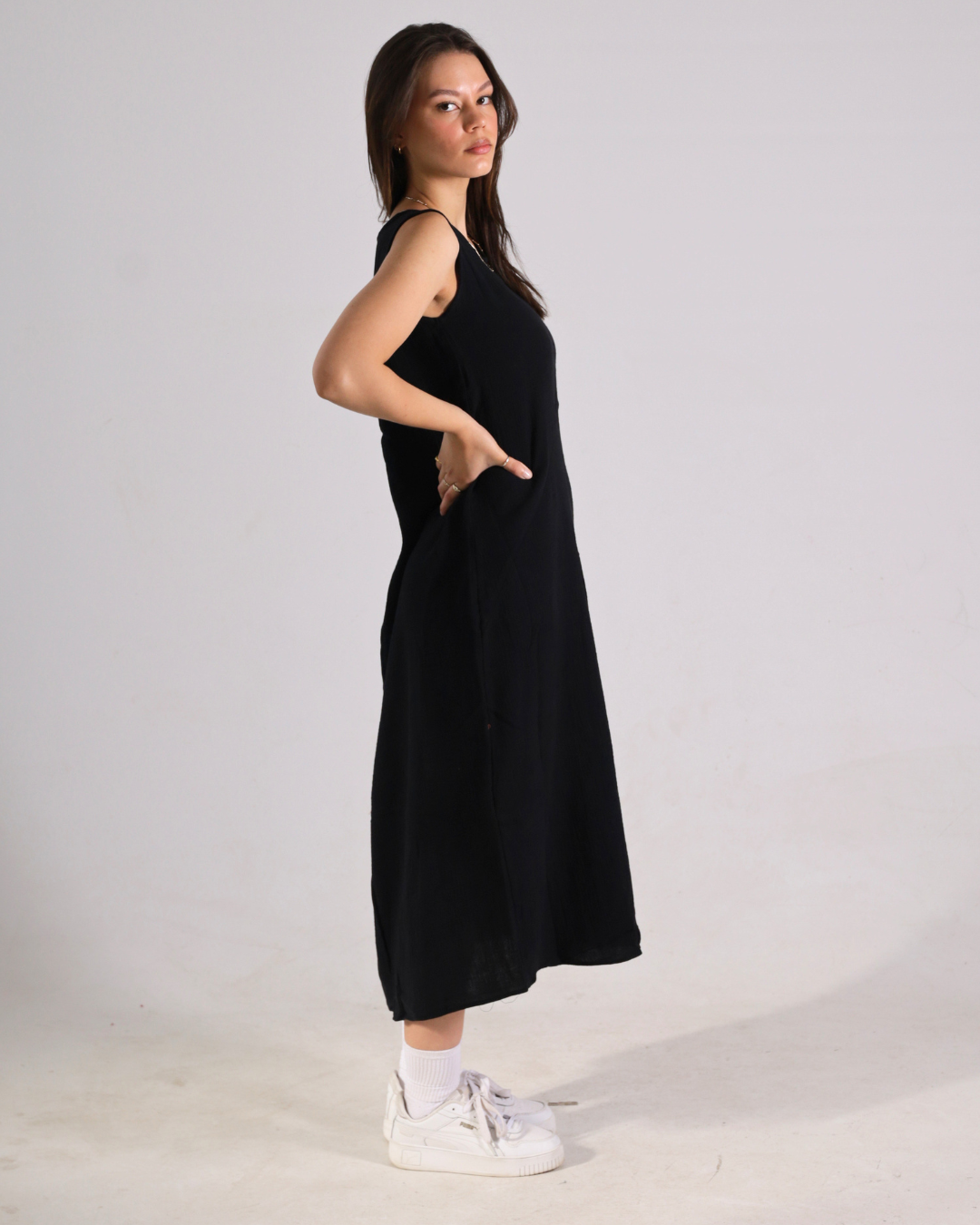 A woman modeling a sleeveless black midi dress with side pockets, paired with white socks and white sneakers, standing against a plain light gray studio background.