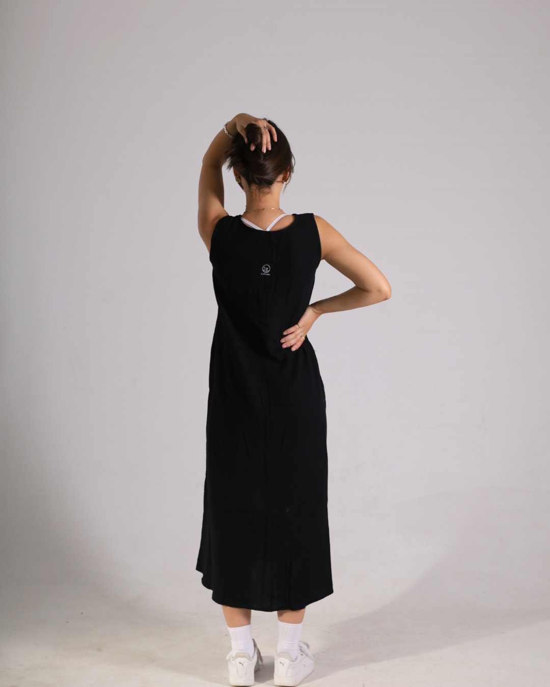 A woman wearing a sleeveless black midi dress, viewed from behind, standing against a plain light gray studio background.