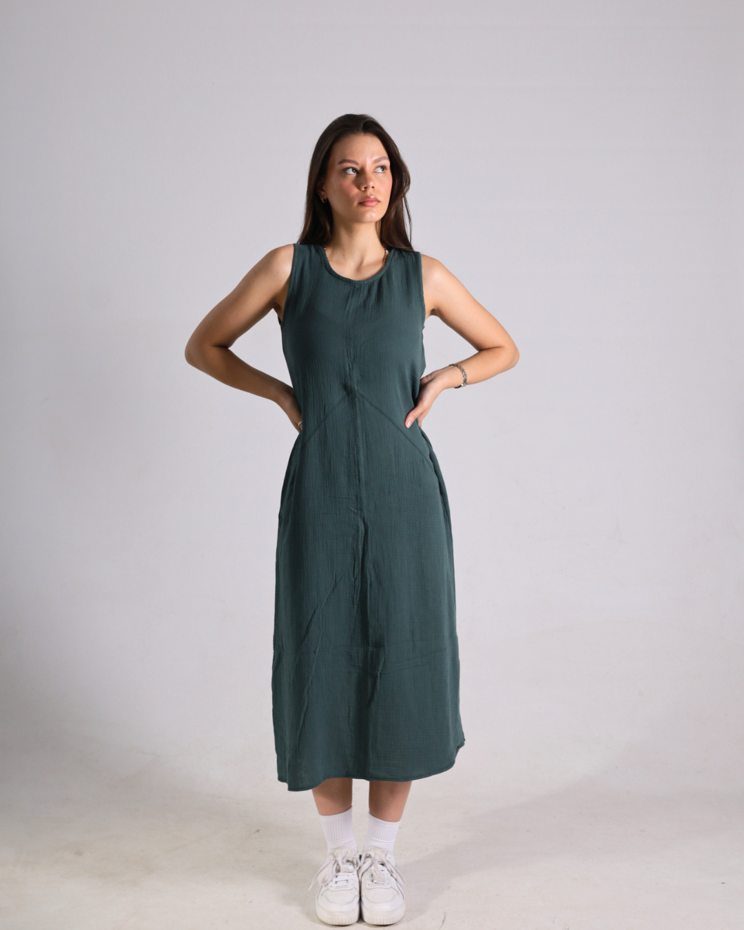 A woman modeling a sleeveless, dark green midi dress with side pockets, paired with white socks and sneakers, standing against a light gray studio background.