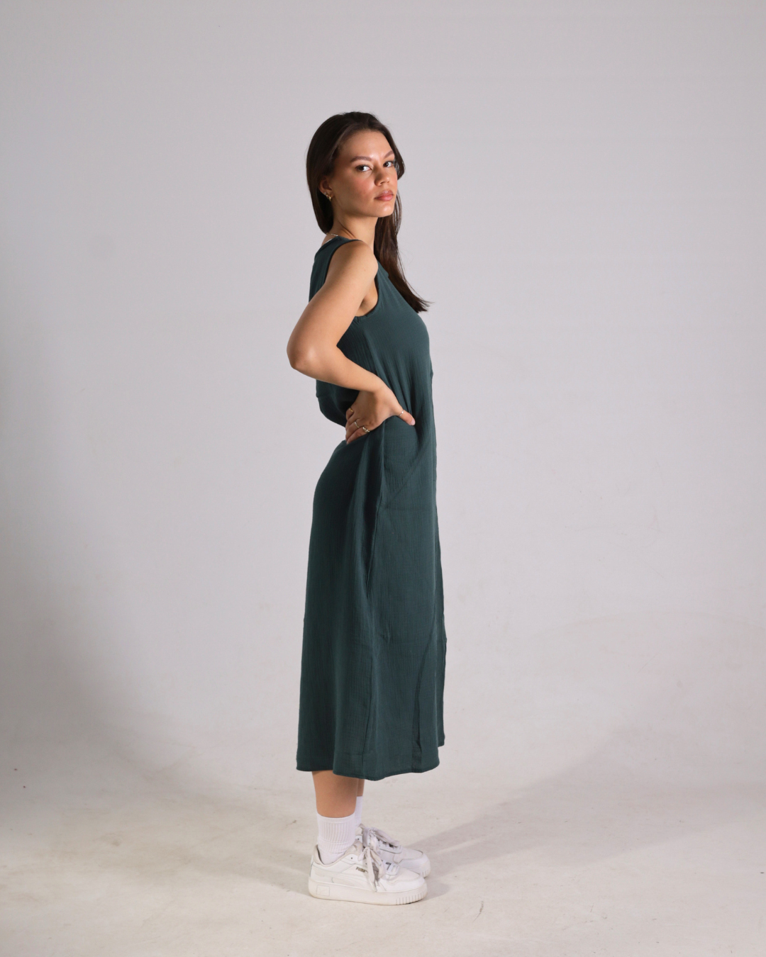 A woman modeling a sleeveless, dark green midi dress with side pockets, paired with white sneakers and socks, standing against a plain light gray studio background.