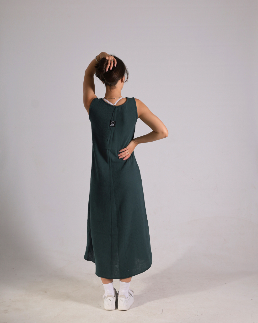 A woman wearing a sleeveless dark green midi dress, shown from the back against a plain light gray studio background.