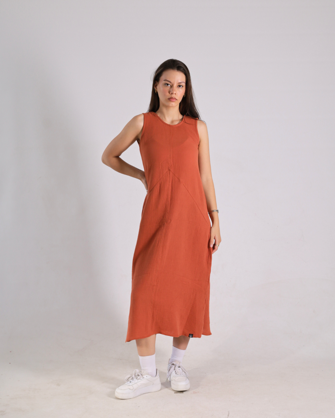 A woman modeling a sleeveless, rust-orange midi dress with a gathered waist, paired with white socks and white sneakers, standing against a light gray studio background.