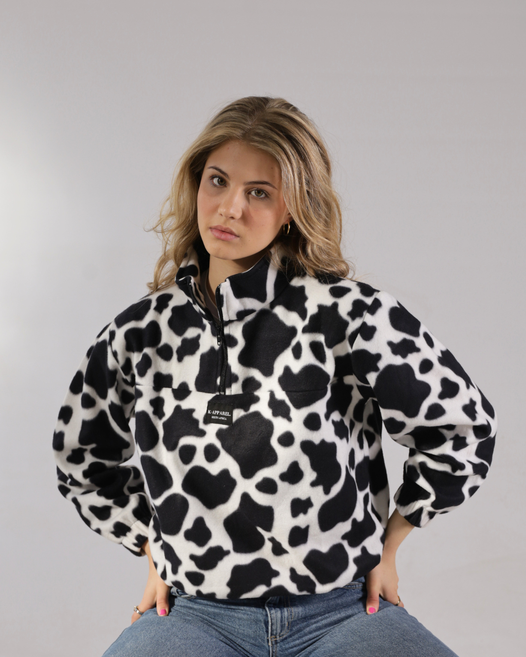 A woman wearing a black and white cow-print fleece pullover jacket with a half-zip collar, posed against a neutral gray studio background.