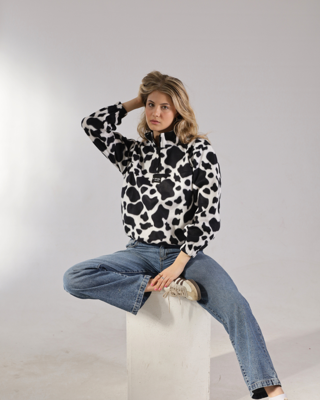 A woman wearing a black and white cow-print fleece half-zip pullover and blue jeans, sitting on a white block against a light gray studio background.