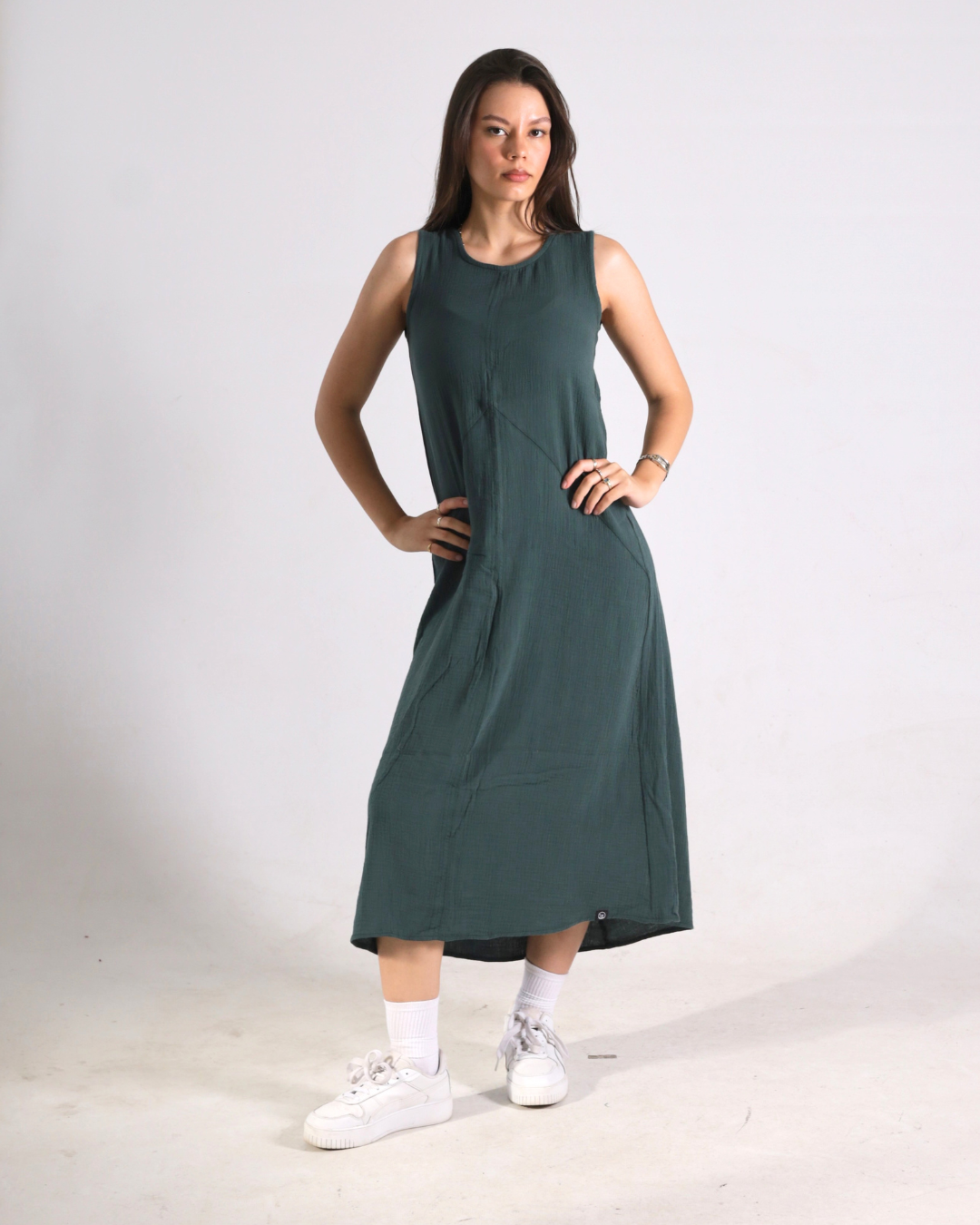 A woman modeling a sleeveless, knee-length dark green dress with white socks and white sneakers, standing against a plain light gray studio background.