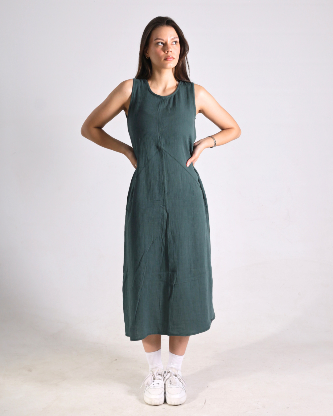 A woman modeling a sleeveless, dark green midi dress with side pockets, paired with white socks and sneakers, standing against a plain light gray studio background.