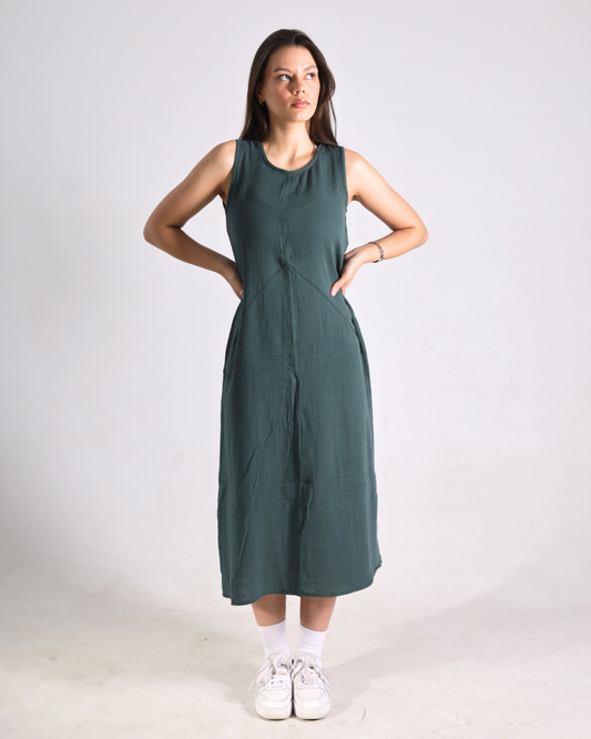 A woman modeling a sleeveless, dark green midi dress with side pockets, paired with white socks and sneakers, standing against a plain light gray studio background.