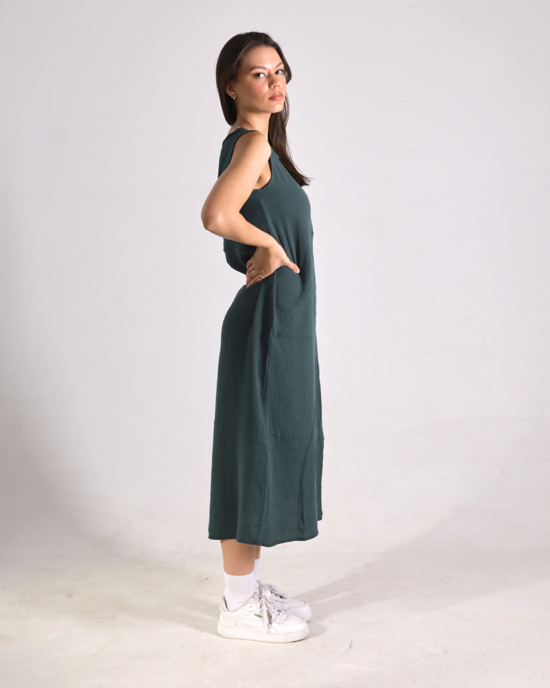 A woman modeling a sleeveless dark green midi dress with side pockets, paired with white socks and white sneakers, standing against a plain light gray studio background.