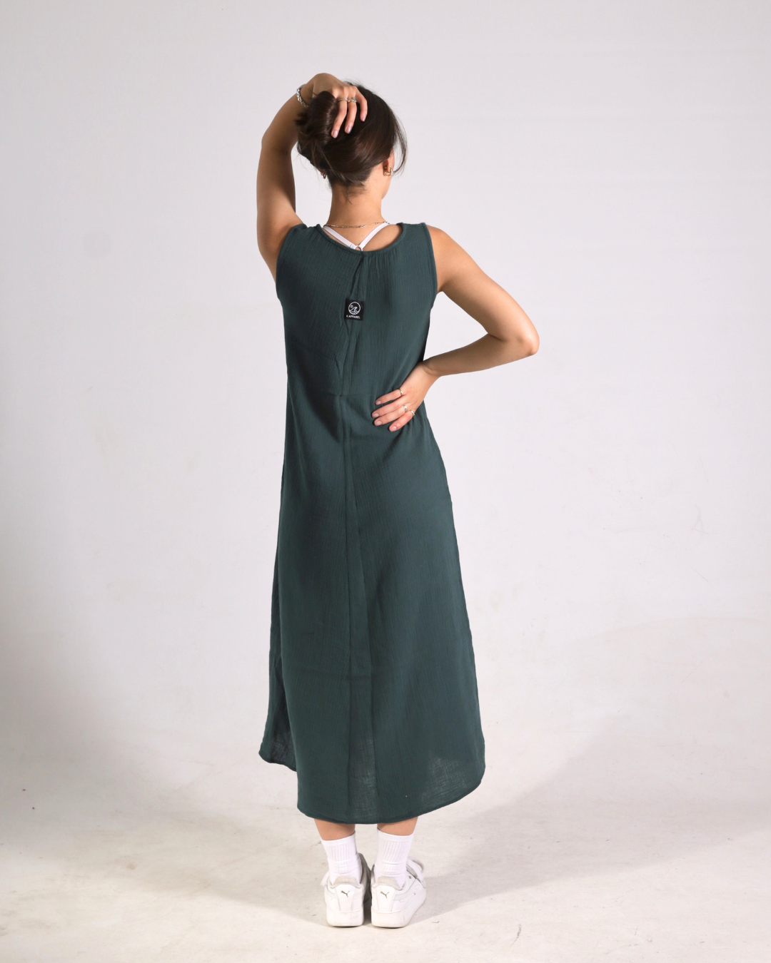 A woman wearing a sleeveless dark green midi dress, viewed from the back, standing against a plain light gray studio background.
