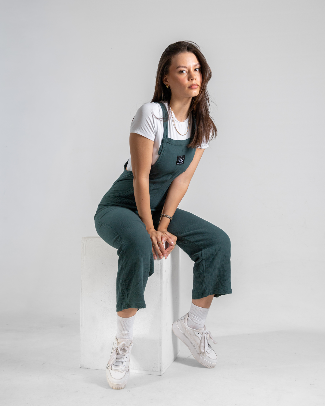 A woman sits on a white cube in a studio setting, wearing dark green overall-style pants over a white t-shirt, paired with white socks and white sneakers.