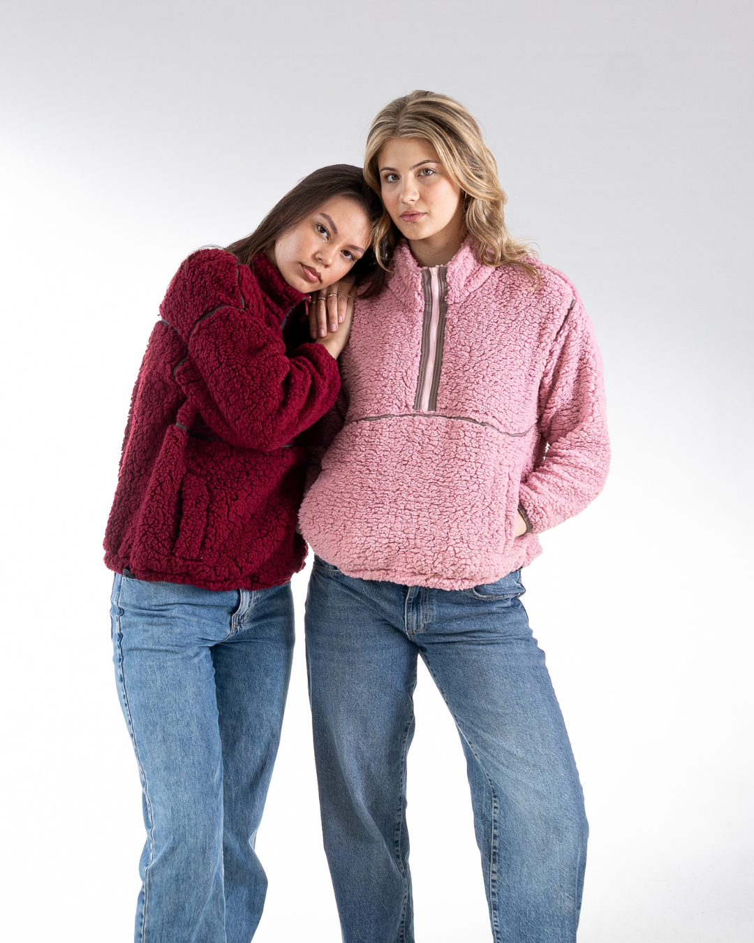 Two women posing together in cozy fleece quarter-zip pullovers, one in deep burgundy and the other in dusty pink, paired with blue jeans against a light gray studio background.