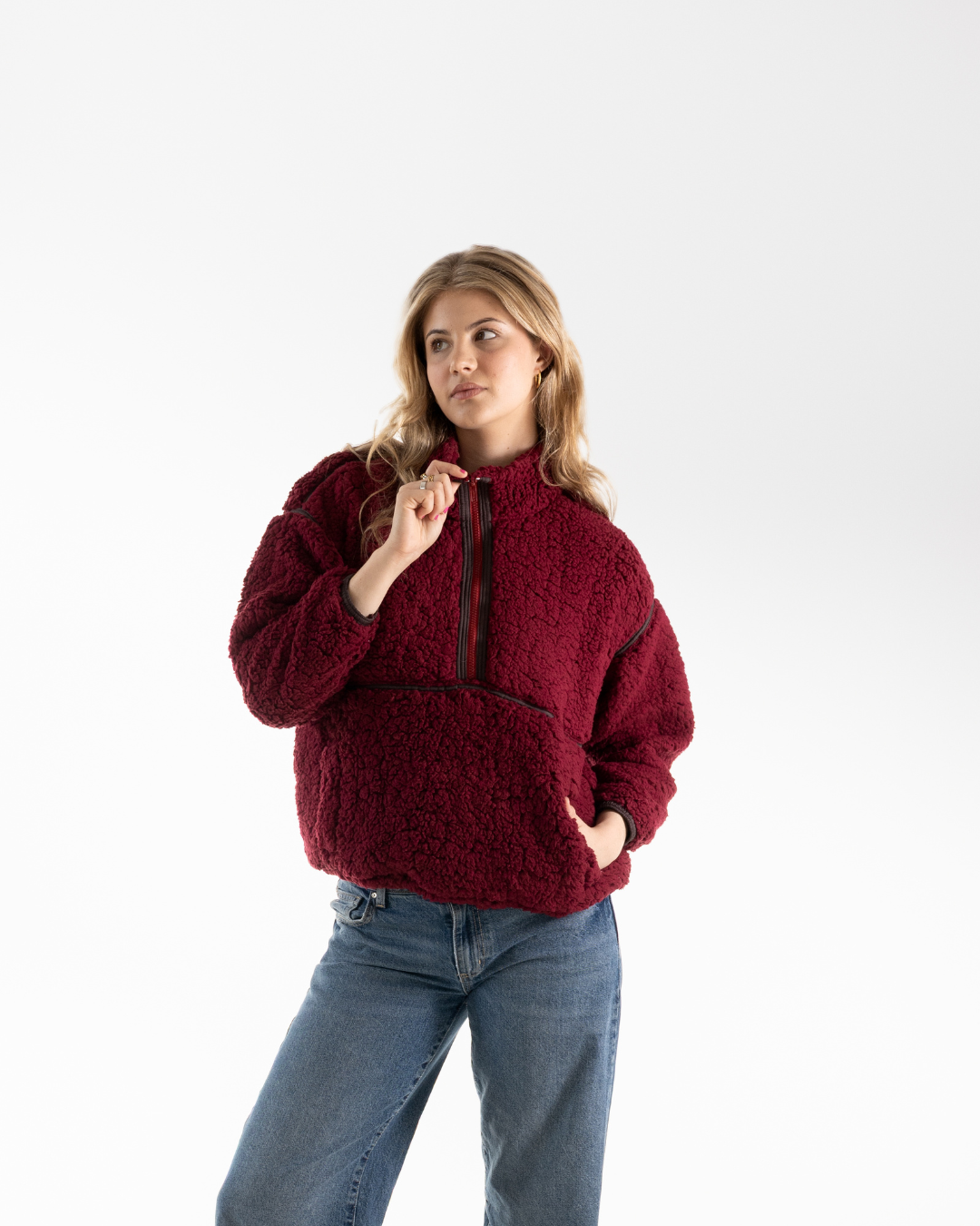 A woman modeling a textured, deep burgundy sherpa fleece half-zip pullover sweater with blue jeans, photographed against a seamless white studio background.