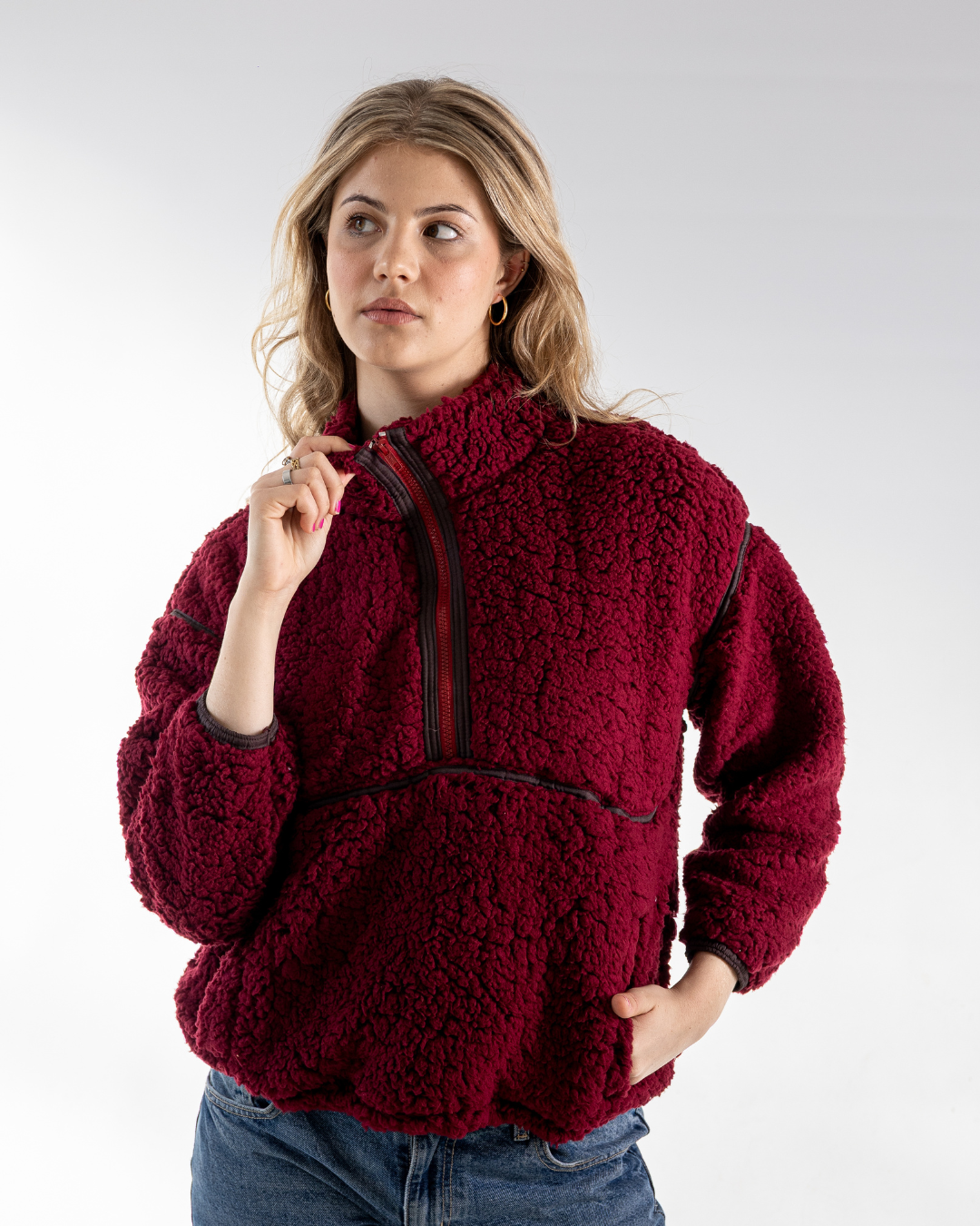 A woman wearing a deep burgundy sherpa fleece quarter-zip pullover jacket with black trim, paired with blue jeans, photographed against a light gray studio background.