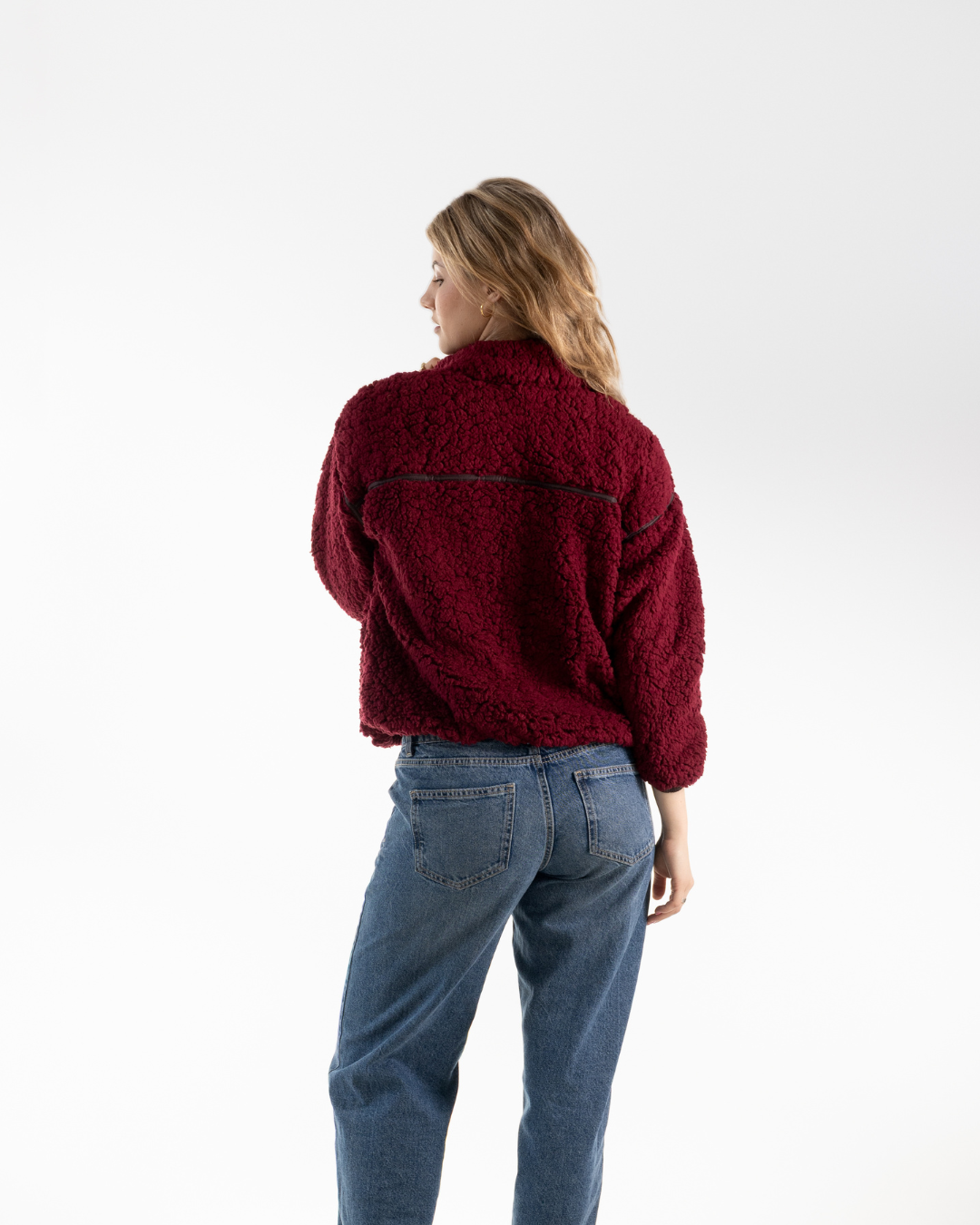 A woman viewed from behind wearing a textured deep burgundy fleece jacket and blue jeans, standing against a plain white studio background.