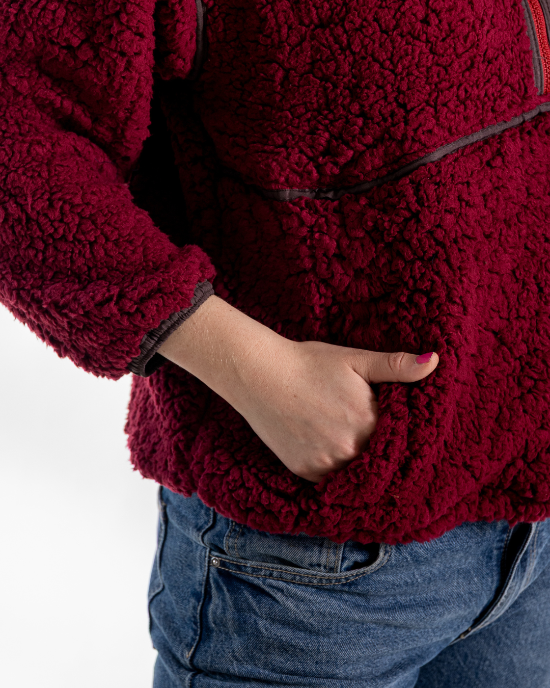 A close-up of a person wearing a deep burgundy sherpa fleece jacket with a zippered pocket and blue jeans, with one hand tucked into the pocket.