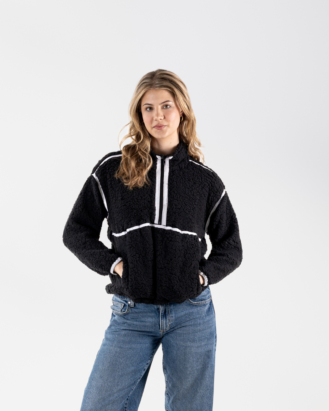 A woman modeling a black sherpa fleece half-zip pullover with white contrast stitching and zipper details, paired with blue jeans, against a plain light gray studio background.