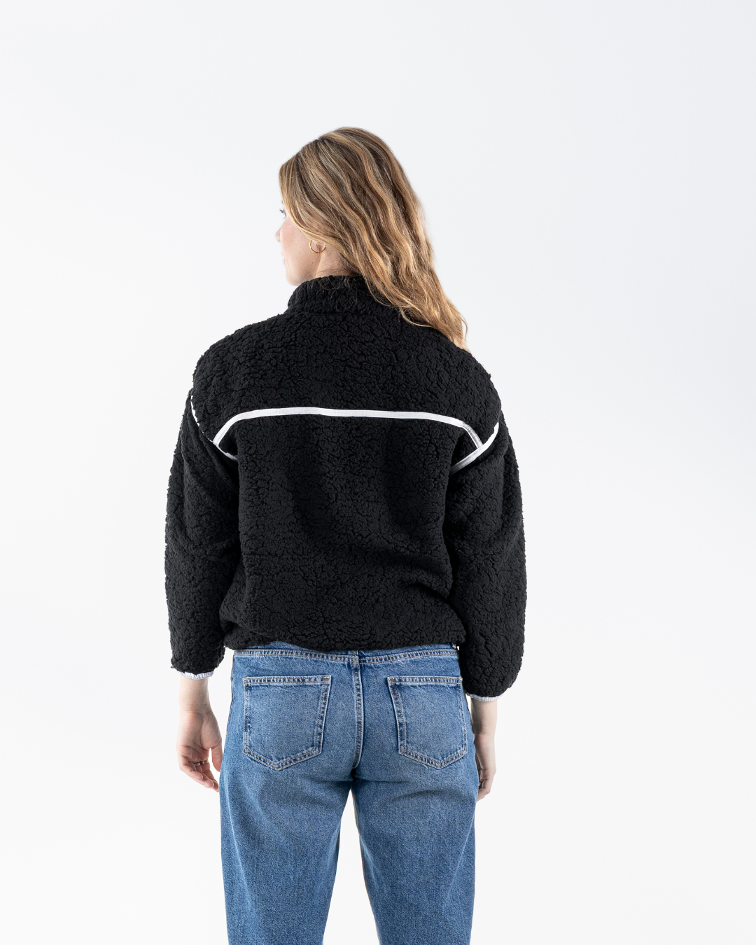 A woman viewed from behind wearing a black textured fleece zip-up jacket with a white horizontal stripe detail across the upper back, paired with blue jeans, against a plain white studio background.