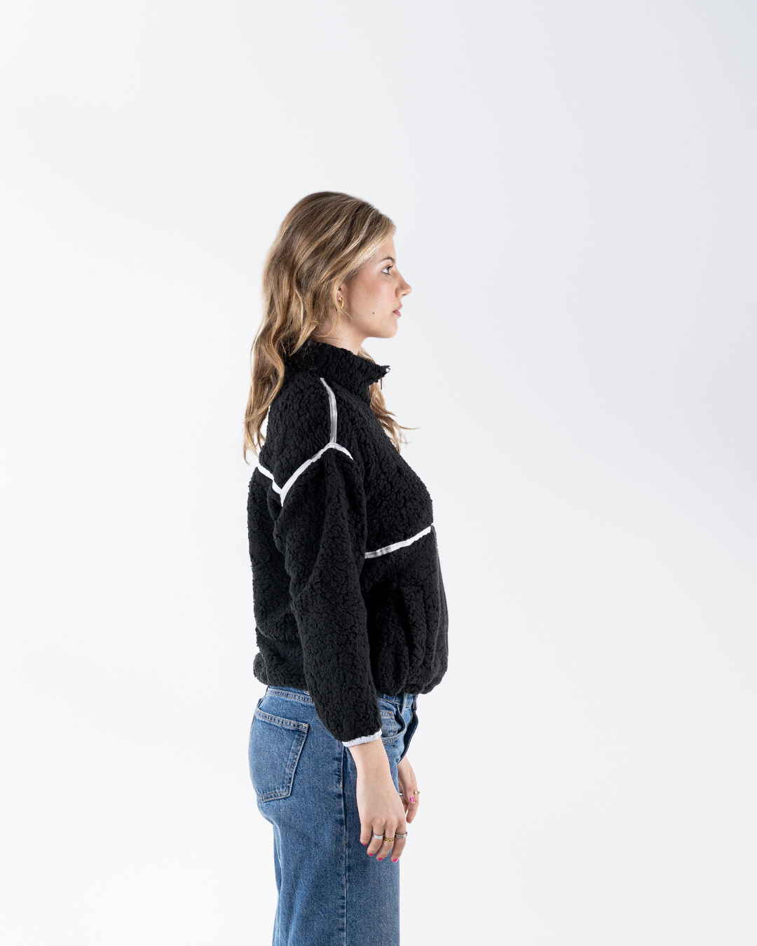 A woman in side profile wearing a black fuzzy cropped sweater with white contrast stitching details and blue jeans, photographed against a clean white studio background.