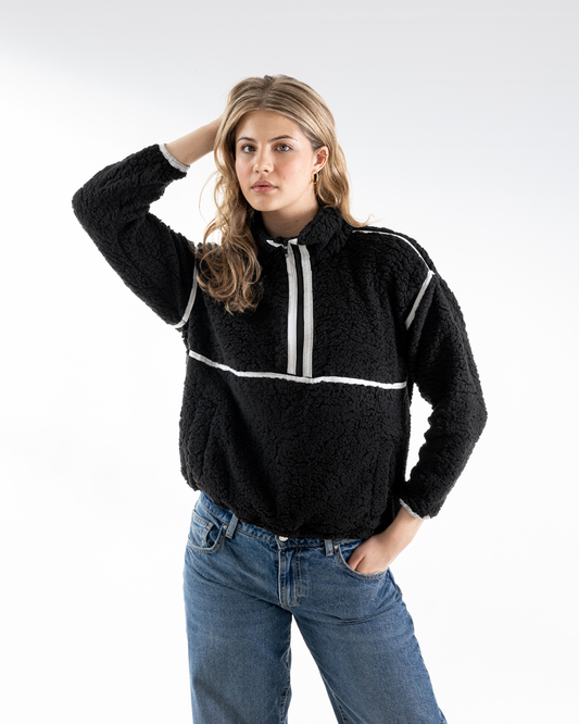 A woman modeling a black textured fleece half-zip pullover sweater with white contrast panels on the chest and shoulders, paired with blue jeans, against a light gray studio background.
