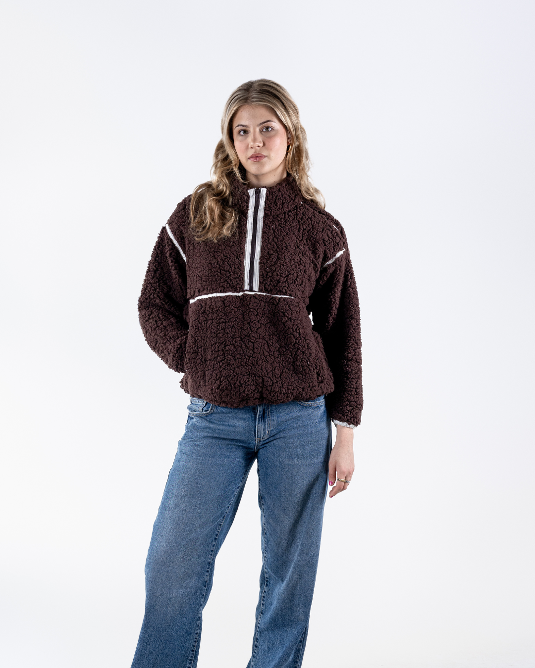 A woman modeling a dark brown textured fleece pullover sweater with white contrast stitching details, paired with blue jeans, against a plain white studio background.