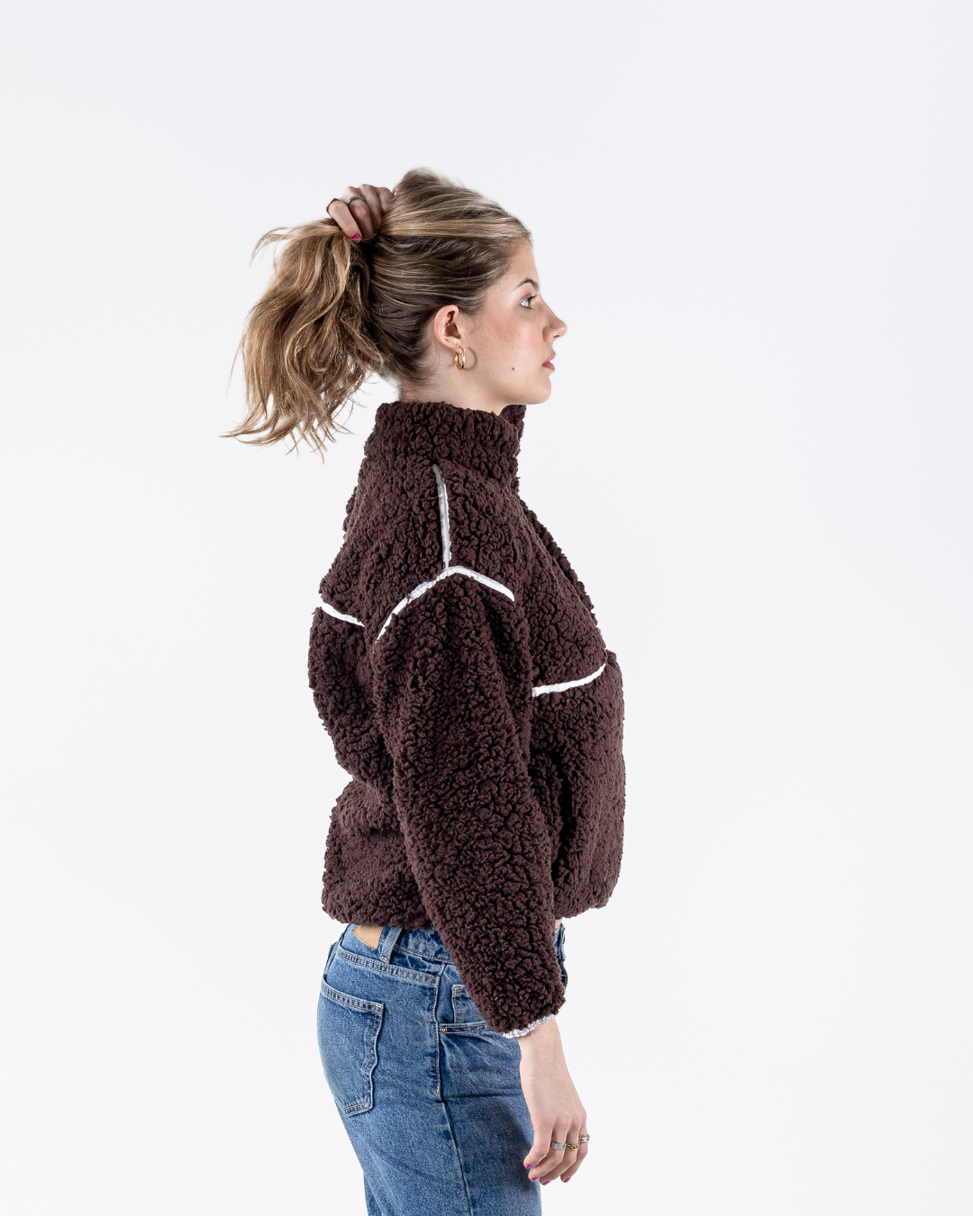 A woman in profile wearing a dark brown sherpa fleece mock-neck sweater with white contrast stitching, paired with blue jeans, against a plain white background.