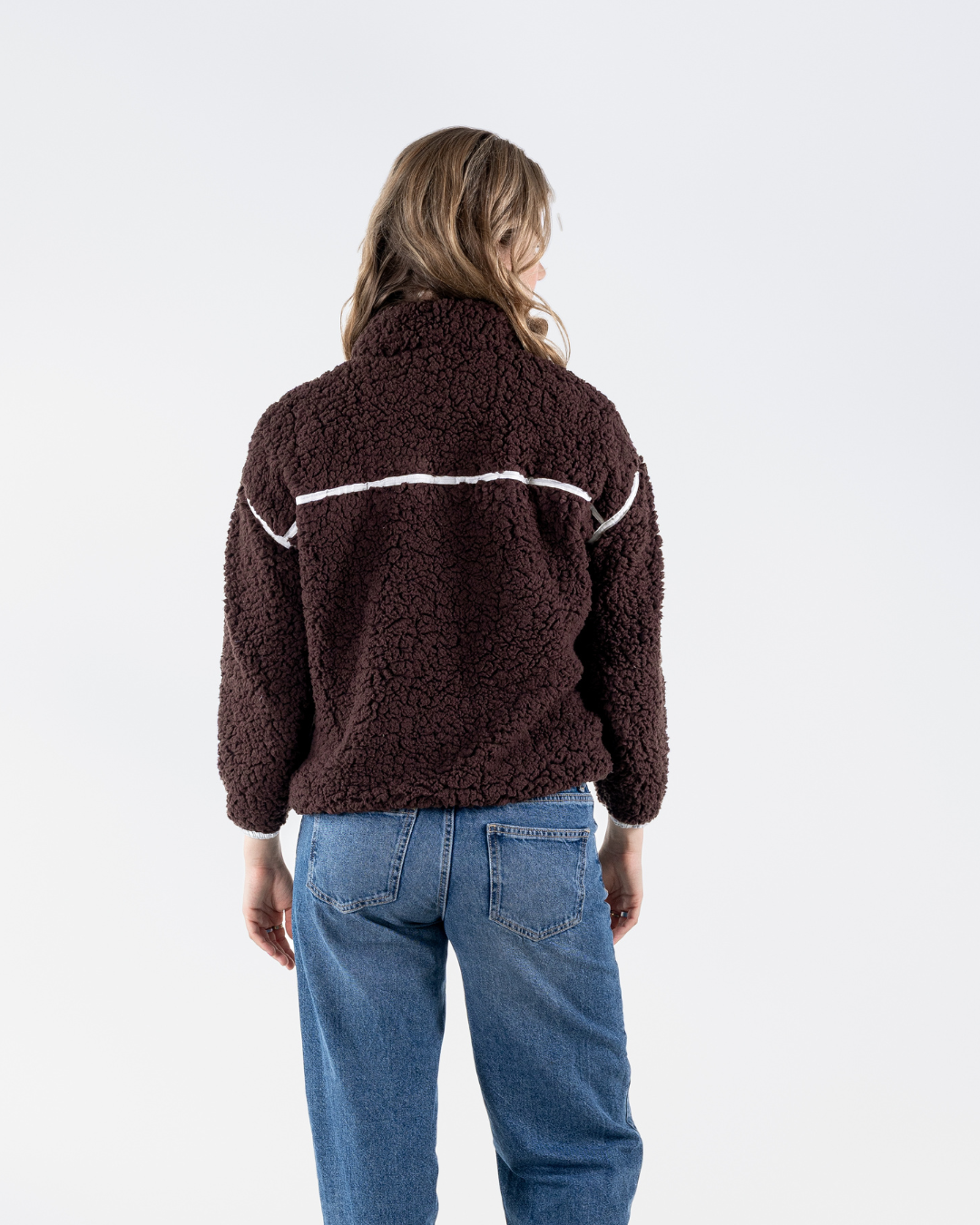 A woman viewed from behind wearing a dark brown sherpa fleece jacket with a white horizontal stripe detail across the upper back, paired with blue jeans, against a plain white studio background.
