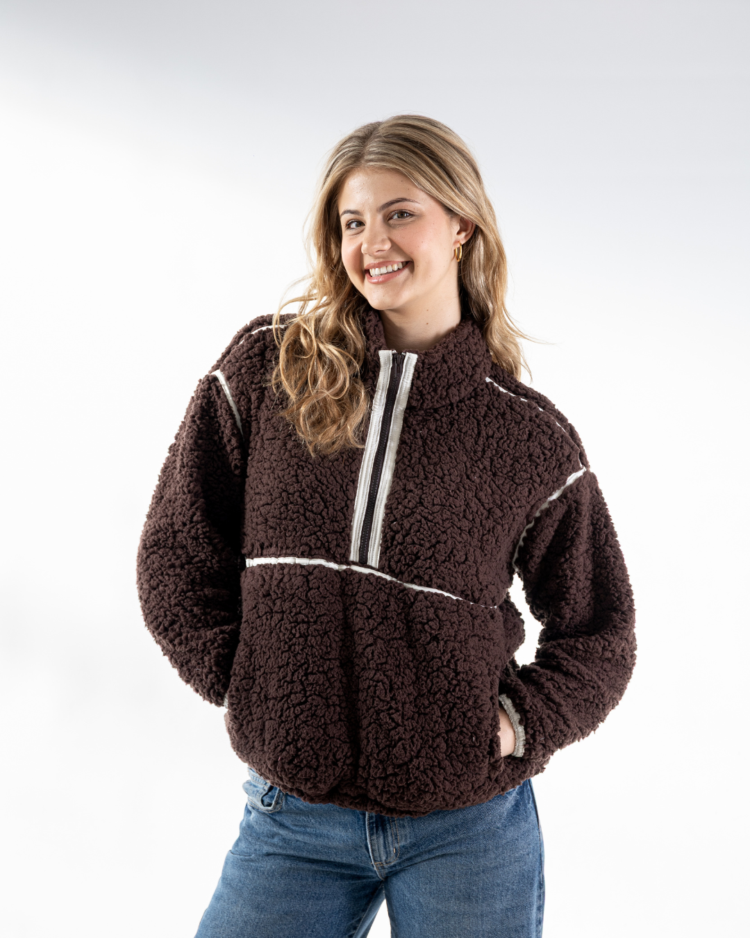 A woman wearing a dark brown sherpa fleece quarter-zip pullover with white contrast stitching and a white zipper trim, paired with blue jeans, against a light gray studio background.