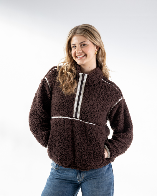 A woman wearing a dark brown sherpa fleece quarter-zip pullover with white contrast stitching and a white zipper trim, paired with blue jeans, against a light gray studio background.