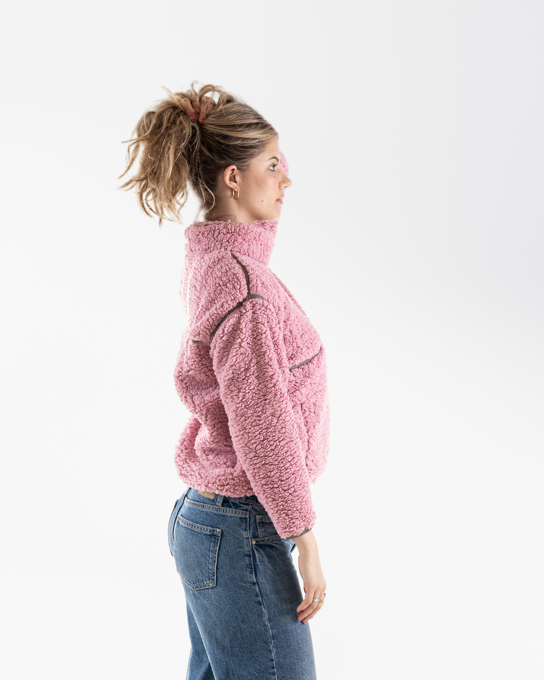 A woman in profile wearing a pink fuzzy cropped fleece jacket and blue jeans, photographed against a clean white studio background.