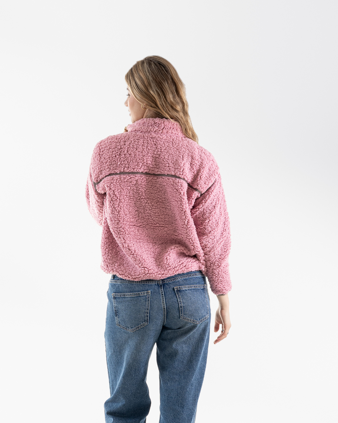 A woman viewed from behind wearing a pink sherpa fleece jacket and blue jeans against a plain white studio background.