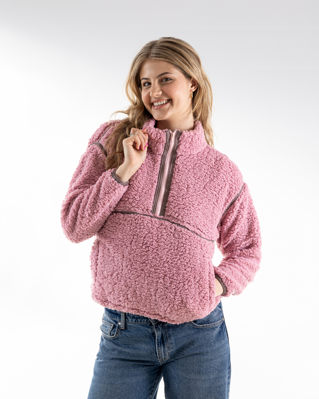 A woman wearing a pink sherpa fleece quarter-zip pullover jacket with gray trim, paired with blue jeans, photographed against a light gray studio background.