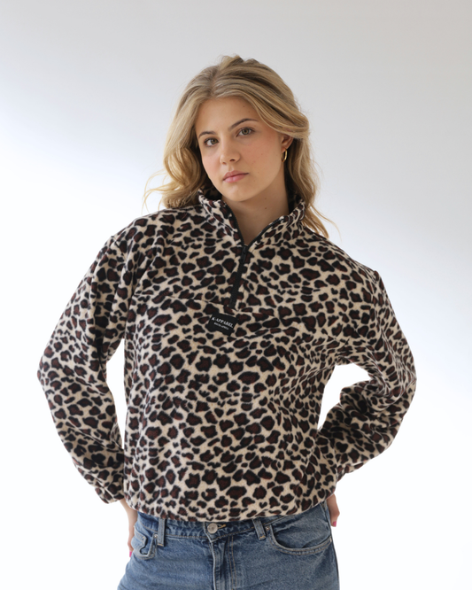 A woman wearing a beige and black leopard print quarter-zip fleece pullover sweater, paired with blue jeans, photographed against a light gray studio background.