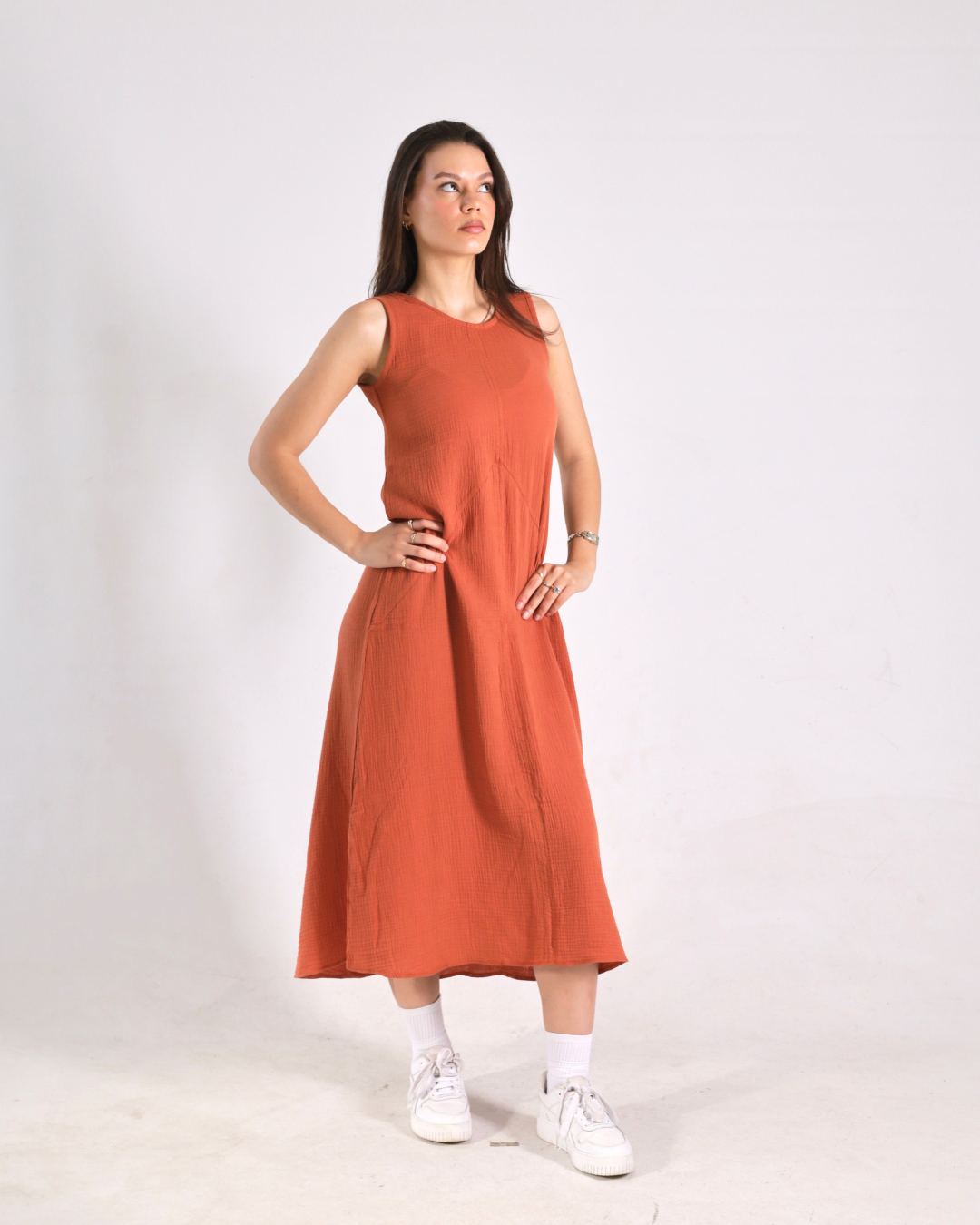 A woman modeling a sleeveless rust-orange midi dress with white socks and white sneakers, standing against a plain light gray studio background.