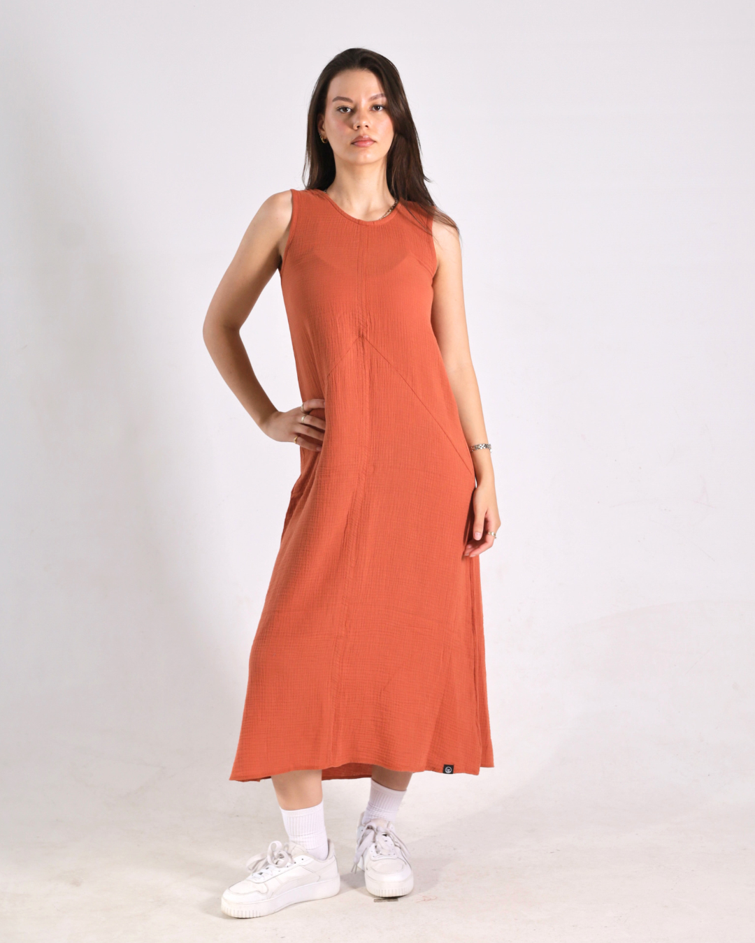 A woman modeling a sleeveless, rust-orange midi dress with diagonal seam details, paired with white socks and white sneakers, against a plain light gray studio background.