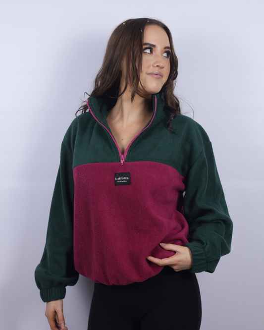 A woman wearing a color-blocked quarter-zip fleece pullover with a dark green upper section and sleeves, and a deep magenta-pink lower front panel. The pullover has a small black brand label on the chest that reads 'A.