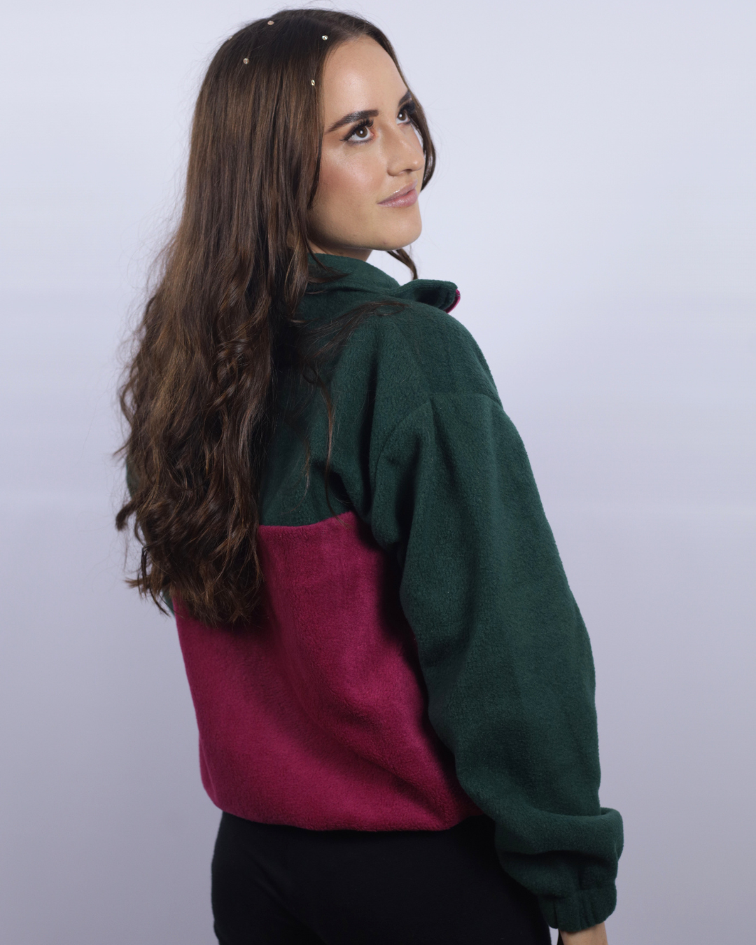 A woman with long brown hair is shown from the back wearing a color-block fleece pullover sweater with dark green and magenta panels, paired with black pants, against a light gray studio background.